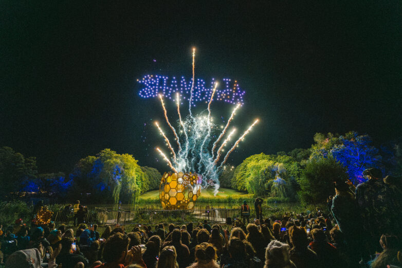 A large crowd watches fireworks and drones forming the word “SHAMBALA” in the night sky above a glowing honeycomb sculpture, with trees and colourful lights.
