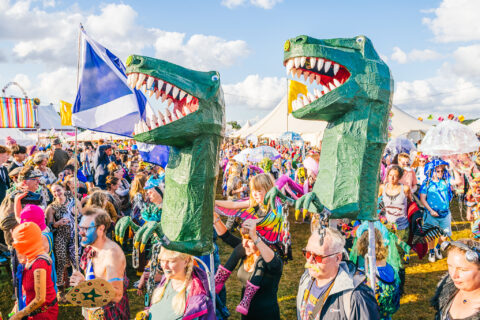A lively outdoor festival crowd, with people in colourful costumes. Two large green dinosaur puppets stand out above the crowd. There are tents, flags, and a bright blue sky with scattered clouds.