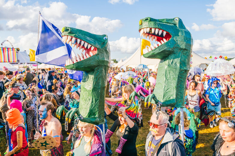 A lively outdoor festival crowd, with people in colourful costumes. Two large green dinosaur puppets stand out above the crowd. There are tents, flags, and a bright blue sky with scattered clouds.