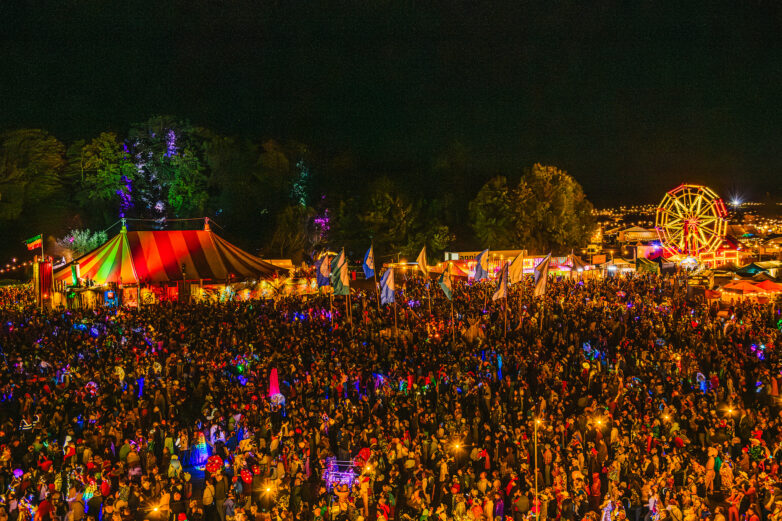 A large, vibrant crowd gathers at a nighttime outdoor festival with colourful lights, a striped circus tent, flags, and a lit Ferris wheel in the background. Trees and more festival activities surround the scene.