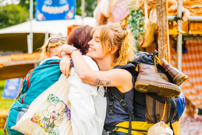 A smiling person with a rucksack and boots hugs another person outdoors, after arriving at Shambala. Both are carrying bags, and the scene is bright and cheerful.