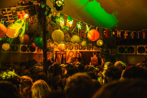 A crowded Back A Yard party with colourful paper lanterns and international flags hanging from the ceiling. People are facing a DJ booth where a DJ is playing music under green and yellow lights.