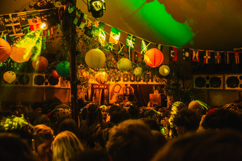 A crowded Back A Yard party with colourful paper lanterns and international flags hanging from the ceiling. People are facing a DJ booth where a DJ is playing music under green and yellow lights.