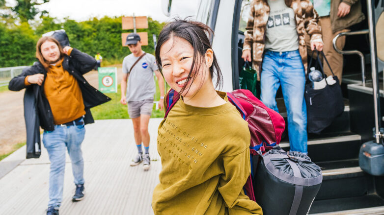 A smiling woman with dark hair holds a sleeping bag and jacket, standing near a coach. Three other people are behind her, carrying bags and rucksacks, outdoors on a bright day.