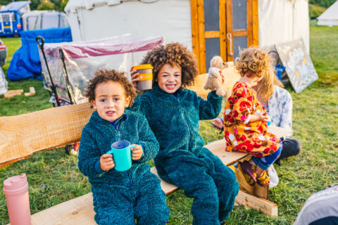 Two young children in fuzzy blue onesies sit on a wooden bench outdoors, smiling and holding cups. A third child in colourful pyjamas sits beside them, holding a teddy bear. A tent and camping gear are visible in the background.