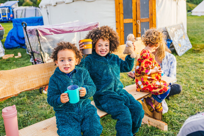 Two young children in fuzzy blue onesies sit on a wooden bench outdoors, smiling and holding cups. A third child in colourful pyjamas sits beside them, holding a teddy bear. A tent and camping gear are visible in the background.
