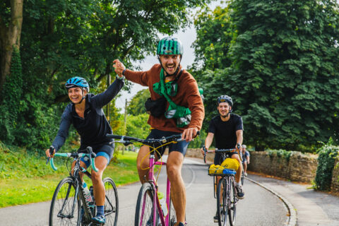 Three adults ride bicycles on a tree-lined road. The two in front, a woman and a man, hold hands and smile cheerfully. A third cyclist follows behind them. All are wearing helmets and casual outfits.
