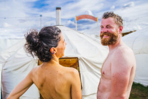 A smiling man and a woman with her back to the camera stand outdoors in front of a white sauna yurt under a partly cloudy sky.