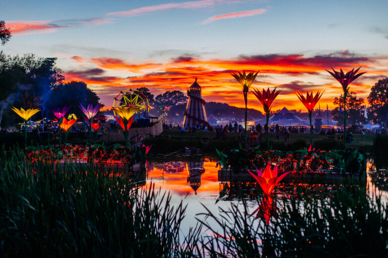 A vibrant festival scene at sunset, featuring glowing, giant flower sculptures by a reflective lake, silhouetted trees, a helter-skelter, and colourful clouds in the sky.