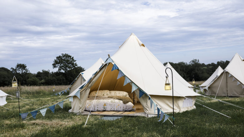 A cosy bell tent set up on green grass, decorated with blue triangular bunting. Other boutique camping tents and lanterns dot the background under a cloudy sky.