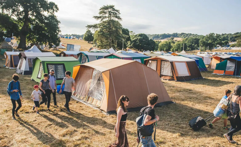People walk among rows of colourful tents set up on a grassy field at a festival campsite, with trees and hills in the background under a partly cloudy sky.