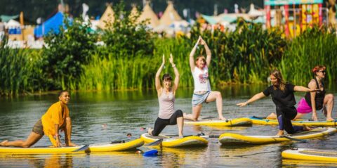 Five people practise yoga poses on paddleboards in a calm, outdoor lake surrounded by green plants and grass. Colourful tents and festival decorations are visible in the blurred background.