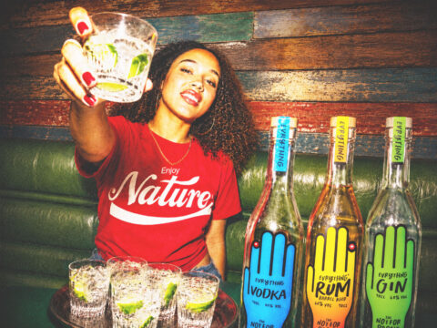 A woman with curly hair and a red Enjoy Nature shirt raises a glass towards the camera, sitting by a table with drinks and three colourful bottles labelled vodka, rum, and gin on a green sofa.