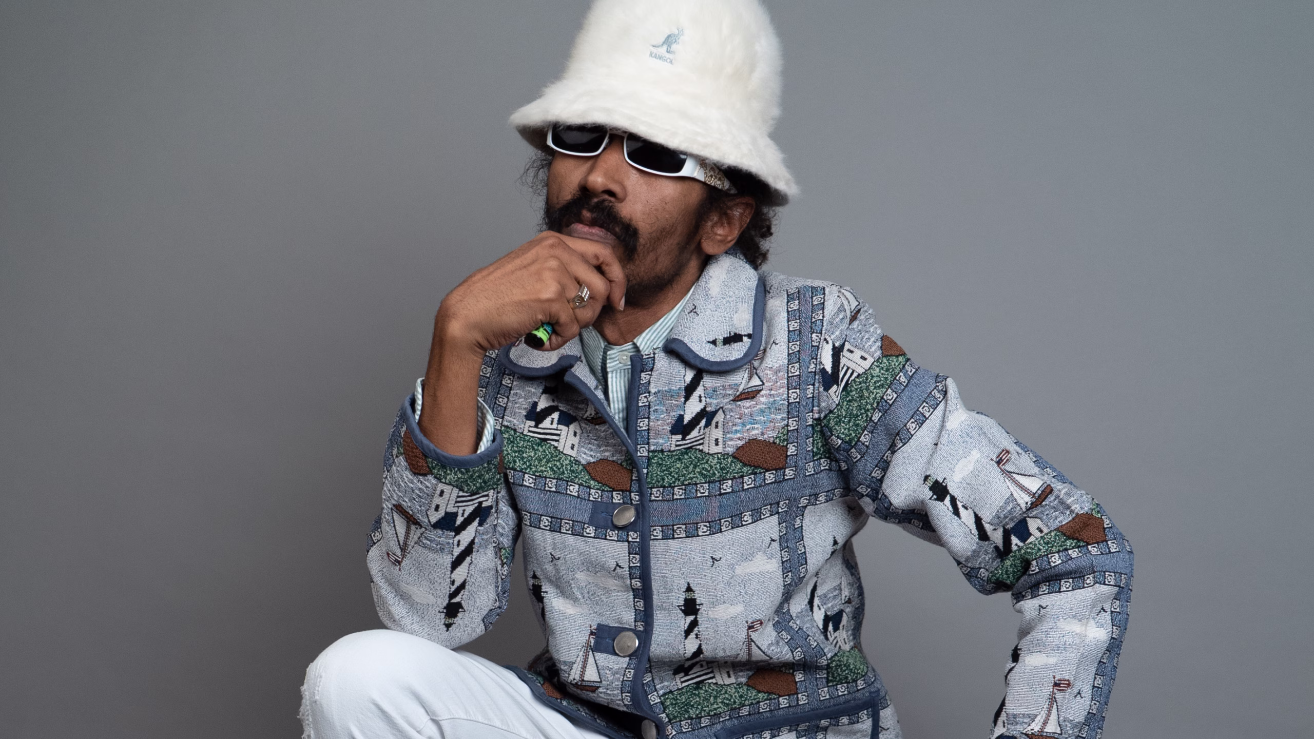 A person wearing a patterned jacket, white trousers, a fuzzy white bucket hat, and white sunglasses poses against a grey background, resting their chin on one hand in a thoughtful manner.
