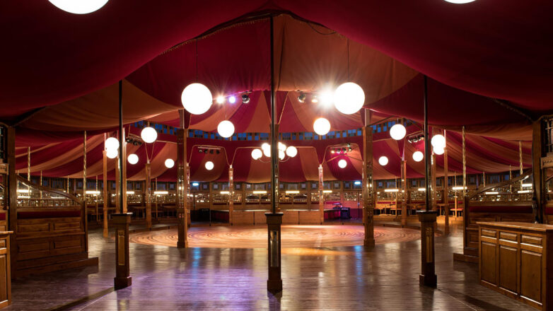 A spacious, circular Spiegeltent with polished wooden floors, warmly lit by globe lights hanging from the red and gold ceiling, surrounded by wooden panelling and mirrors, creating an inviting, vintage atmosphere.