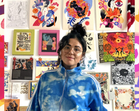 A person wearing glasses and a blue tie-dye Adidas jacket stands in front of a wall covered with colourful, artistic prints and illustrations.